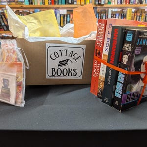 May include: A brown box with the text "COTTAGE used BOOKS" on the front. The box is filled with books wrapped in orange ribbon and a clear bag with bookmarks. The books are titled "Wild Storm", "Open Season", "Blood Hunt", "Ian Rankin", "C.J. Box", and "Richard Castle".