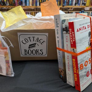 May include: A brown cardboard box labeled Cottage Used Books filled with yellow and orange tissue paper. Beside the box sits a stack of three books tied with an orange ribbon, and a small sheer white drawstring bag containing paper items.
