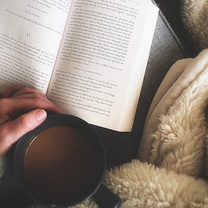 May include: A person is reading a book while holding a mug of coffee. The person is sitting on a couch with a fluffy blanket.
