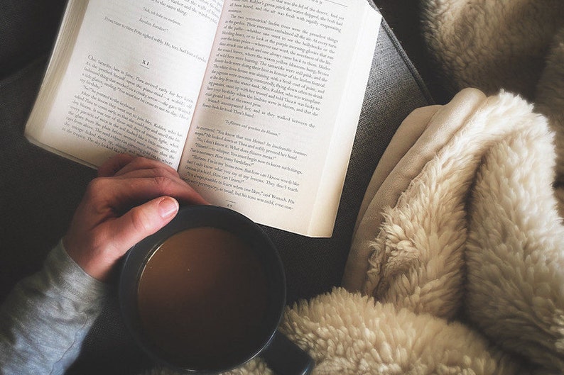 May include: A person is reading a book while holding a mug of coffee. The person is sitting on a couch with a fluffy blanket.
