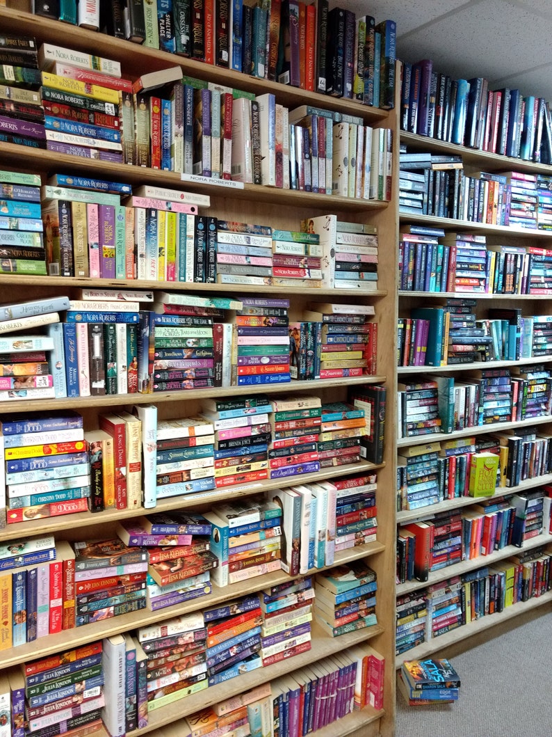 May include: Two tall wooden bookshelves filled with a large collection of books arranged in multiple rows. The shelves contain various hardcover and paperback novels with colorful spines, some stacked horizontally and others standing vertically.