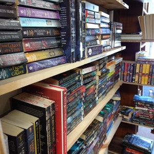 May include: Multiple wooden bookshelves filled with various paperback and hardcover books. The collection includes titles by authors such as Jacquelyn Frank, Sherrilyn Kenyon, and Robin McKinley, organized on shelves with a label reading Paranormal Romance.