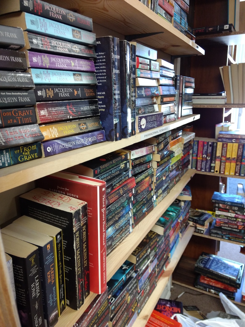 May include: Multiple wooden bookshelves filled with various paperback and hardcover books. The collection includes titles by authors such as Jacquelyn Frank, Sherrilyn Kenyon, and Robin McKinley, organized on shelves with a label reading Paranormal Romance.