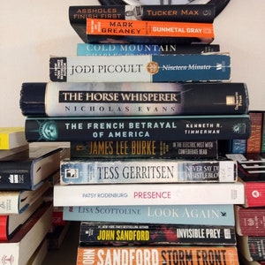 May include: A stack of paperback books on a shelf. The titles include "Assholes Finish First", "Cold Mountain", "The Horse Whisperer", "The French Betrayal", "Tess Gerritsen", "Presence", "Look Again", "Invisible Prey", "Storm Front", and "Misery Bay".