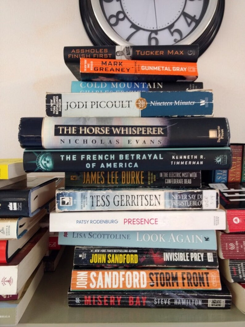 May include: A stack of paperback books on a shelf. The titles include "Assholes Finish First", "Cold Mountain", "The Horse Whisperer", "The French Betrayal", "Tess Gerritsen", "Presence", "Look Again", "Invisible Prey", "Storm Front", and "Misery Bay".