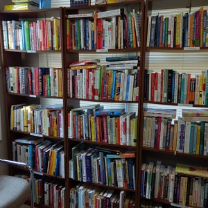 May include: A full bookshelf with multiple rows of books, mostly in the self-help and business genres. The books are organized by color and size, and there are many different titles and authors represented. The bookshelf is made of wood and has a light brown finish.