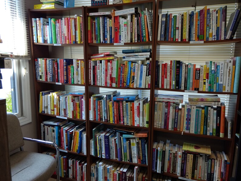 May include: A full bookshelf with multiple rows of books, mostly in the self-help and business genres. The books are organized by color and size, and there are many different titles and authors represented. The bookshelf is made of wood and has a light brown finish.