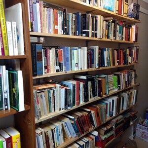 May include: A tall wooden bookshelf unit featuring multiple horizontal shelves filled with a large collection of books of varying sizes, colors, and thicknesses, arranged in vertical rows with spines facing outward for easy identification.