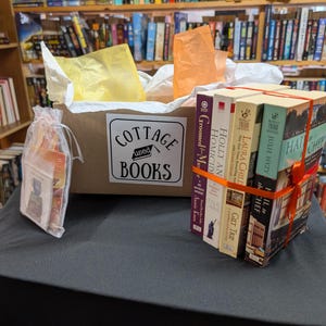 May include: A cardboard box labeled "COTTAGE USED BOOKS" filled with books and tissue paper. A bundle of books tied with orange ribbon and a small gift bag are on a black surface. Bookshelves are in the background.