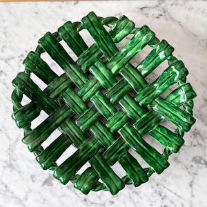 May include: A round, deep green ceramic basket featuring a woven lattice design. The surface has a glossy, reflective finish with variations in color intensity across the interconnected strips, creating a textured, open-work pattern throughout.