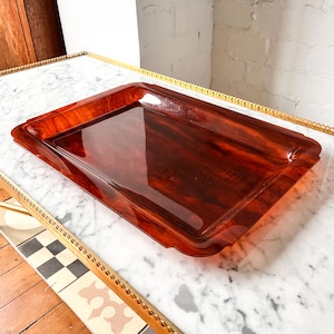 May include: A rectangular, reddish-brown serving tray with a glossy finish. The tray has a slightly raised edge and a decorative, scalloped design along the rim. The tray is made of a material that resembles tortoiseshell.