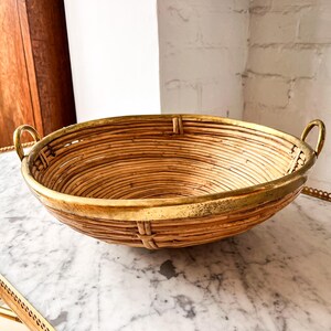 May include: A round, woven rattan basket with a gold-coloured metal rim and handles. The basket is light brown with a textured surface and is sitting on a marble surface.