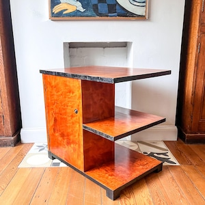May include: A wooden side table with a cabinet door and two shelves. The table is made of light wood with a dark wood trim. The table is in a room with a white wall and a fireplace.