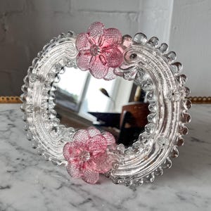 May include: A clear glass mirror with a decorative frame. The frame features a scalloped edge and is adorned with pink glass flower accents. The mirror is round and reflects the surrounding environment. The overall design is delicate and ornate.