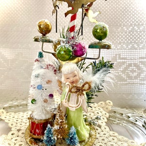 May include: A vintage Christmas centerpiece featuring a ceramic angel in a green dress holding a harp. The centerpiece includes a white bottle brush tree, red drum, and gold-colored metal accents with ornaments and a candy cane.