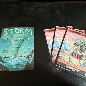 May include: Collectible trading cards with fantasy illustrations. One card shows a turquoise tornado with the text "STORM COUNTER". Three cards depict a green goblin with a bomb in a mountain landscape.