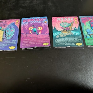 May include: Four fantasy trading cards with vibrant illustrations and text. The cards, in pink, blue, and green, feature creatures and are labelled "Goblin Recruiter", "Goblin Lackey", "Muxus", and "Goblin Matron". The cards are displayed on a dark surface.