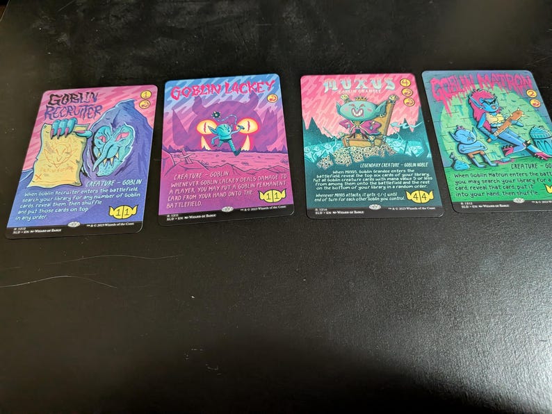 May include: Four fantasy trading cards with vibrant illustrations and text. The cards, in pink, blue, and green, feature creatures and are labelled "Goblin Recruiter", "Goblin Lackey", "Muxus", and "Goblin Matron". The cards are displayed on a dark surface.