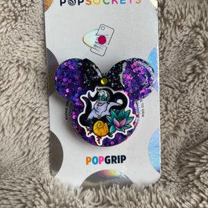 May include: A purple glitter PopSockets PopGrip in the shape of Mickey Mouse ears. The center features a graphic of Ursula from The Little Mermaid. The PopSockets packaging is visible in the background.