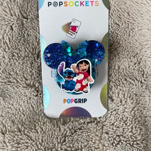 May include: A blue and pink glitter phone grip featuring Stitch and Lilo from Disney's Lilo & Stitch. The grip is shaped like Mickey Mouse ears and has the words "PopGrip" printed on the bottom.