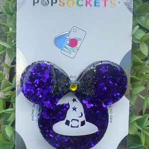 May include: A purple glitter PopSocket phone grip in a mouse ear shape, with a black bow and a white witch hat design. The PopSocket is attached to a white backing with the PopSockets and PopGrip logos.