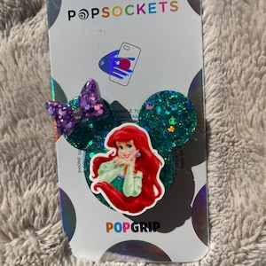 May include: A PopSockets PopGrip featuring a green glitter background with a purple glitter bow and a sticker of Ariel from The Little Mermaid. The text "PopGrip" is printed in white on the bottom of the card.