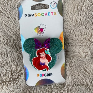May include: A green glitter phone grip with a purple bow and a sticker of Ariel from The Little Mermaid. The grip is packaged on a white card with the words "PopSockets" and "PopGrip".
