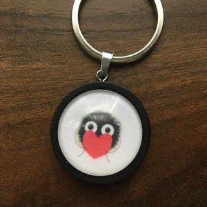 May include: A black and white round keychain with a cartoon image of a creature holding a red heart. The keychain has a silver metal ring.