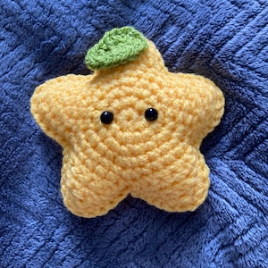 Könnte beinhalten: Ein gelber Häkelstern mit einem grünen Blatt oben. Der Stern hat schwarze Augen und einen lächelnden Mund.