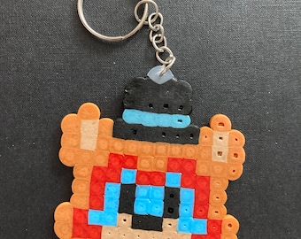 May include: A brown bear-shaped keychain made of perler beads. The bear is wearing a black hat with a blue brim. The keychain has a silver metal ring.