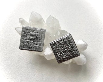 May include: A pair of square stud earrings featuring a metallic silver finish with a textured, crinkled surface pattern. The earrings are positioned resting against a cluster of translucent, pointed white quartz crystal formations.