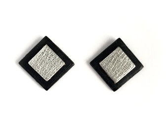 May include: A pair of square stud earrings featuring a black outer border and a textured silver-colored center square. The design is geometric and flat, with the silver square layered on top of the black square base to create a two-tone contrast.