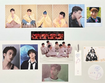 May include: A collection of BTS merchandise, including photo cards, a film strip, a bookmark, and a sticker. The photo cards feature individual members and group shots. The items are arranged on a white surface.