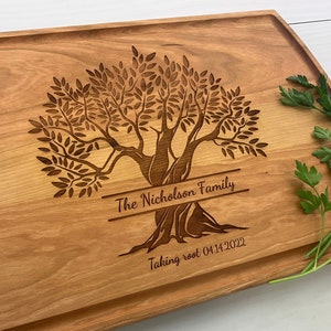 May include: A wooden cutting board with a carved olive tree design. The board is engraved with "The Nicholson Family" and "Taking root 04.14.2022".