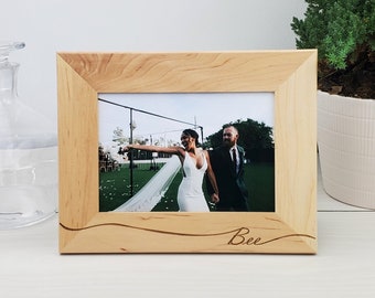 May include: A wooden picture frame with a light brown finish. The frame has a wavy design at the bottom and the word "Bee" is engraved in the wood. The frame contains a photo of a bride and groom.