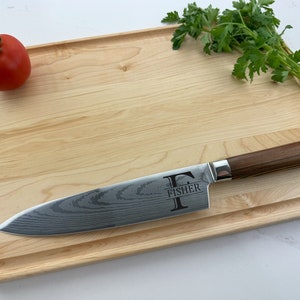 May include: A personalized maple wood cutting board with a  knife on top. The knife has a brown handle and a stainless steel blade with a damascus pattern. The cutting board has a rounded edge and a groove around the edge. The knife has the letter "F" and the name "Fisher" engraved on the blade.