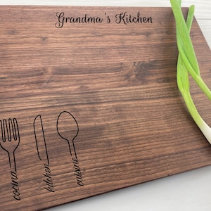 Peut inclure: Une planche à découper en bois brun avec le texte "Grandma's Kitchen" gravé sur le dessus. La planche comporte trois images stylisées d'une fourchette, d'un couteau et d'une cuillère avec les mots "cucina", "kitchen" et "cuisine" écrits sous chaque image.