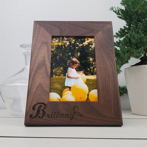 May include: A dark brown wooden picture frame with a personalized inscription "Brittany" and a photo of a young girl playing with yellow balloons.