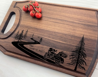 May include: A rectangular wooden cutting board with a handle, featuring a carved design of a motorcycle on a winding road with trees. The text "Jimmy... Adventure Awaits" is included. A cluster of red cherry tomatoes rests on the board.