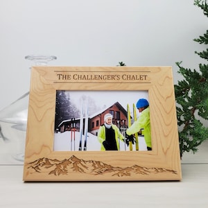 May include: A wooden picture frame with a photo of two people skiing in front of a chalet. The frame is engraved with the text "The Challenger's Chalet" and a mountain range.