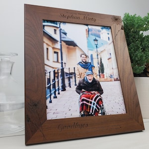 May include: A wooden picture frame with a photo of two people walking down a street. The frame has a floral design and the text "In Loving Memory of Stephan Minty" and "Goodbyes are not forever - goodbye's are not the end - they simply mean I'll miss you until we meet again."