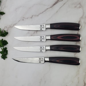 May include: Four steak knives with serrated blades and dark wood handles. The blades have a patterned design and the word "FISHER" is printed on the blade. The handles are a dark brown with red accents.