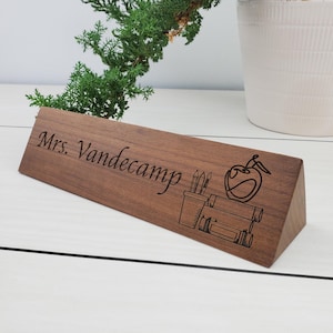 May include: A wooden desk nameplate with the text "Mrs. Vandecamp" engraved on it. The nameplate is triangular in shape and features a design of an apple, pencils, and a ruler.