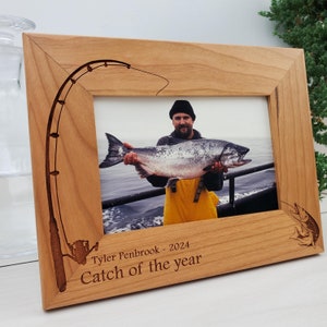 Peut inclure: Un cadre photo en bois avec un motif de canne à pêche gravé. Le cadre contient une photo d'une personne tenant un gros poisson. Le texte "Tyler Penbrook - 2024 Catch of the year" est gravé sur le cadre.