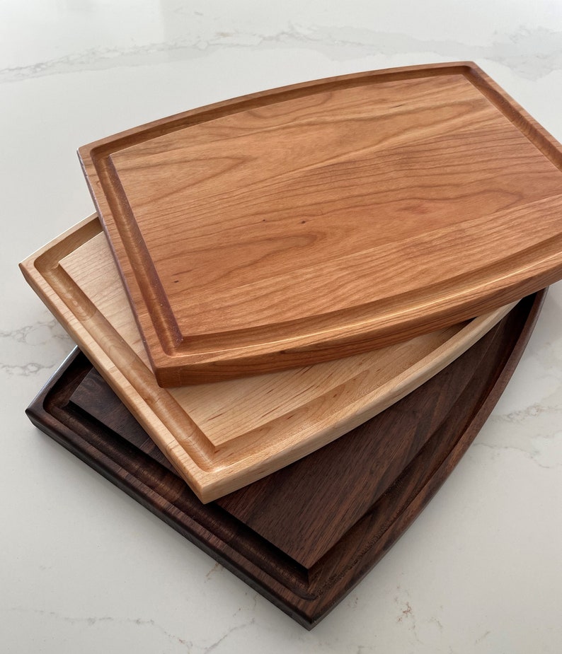 May include: Three wooden cutting boards stacked on top of each other. The top board is cherry wood, the middle board is maple wood, and the bottom board is walnut wood. All three boards have a rounded edge.