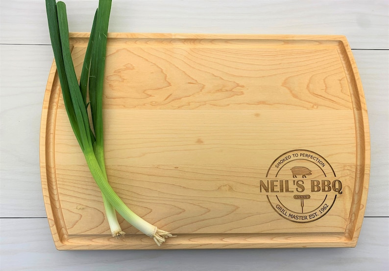 May include: A large, rectangular wooden cutting board with rounded edges. The board is made of light wood and has a natural grain pattern. The board is engraved with the text "Smoked to Perfection Neil's BBQ Grill Master Est. 1952" in a circle with a pig illustration.