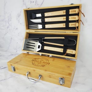 May include: A bamboo BBQ tool set in an open wooden case. The set includes a spatula, tongs, knife, fork, and basting brush. The case is engraved with "Dave's BBQ" and has a handle and metal clasps.