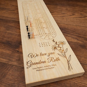 May include: A wooden cribbage board with a floral design and the text "We love you Grandma Ruth". Includes metal pegs in black, silver, and copper colors. The board is made of light-colored wood.