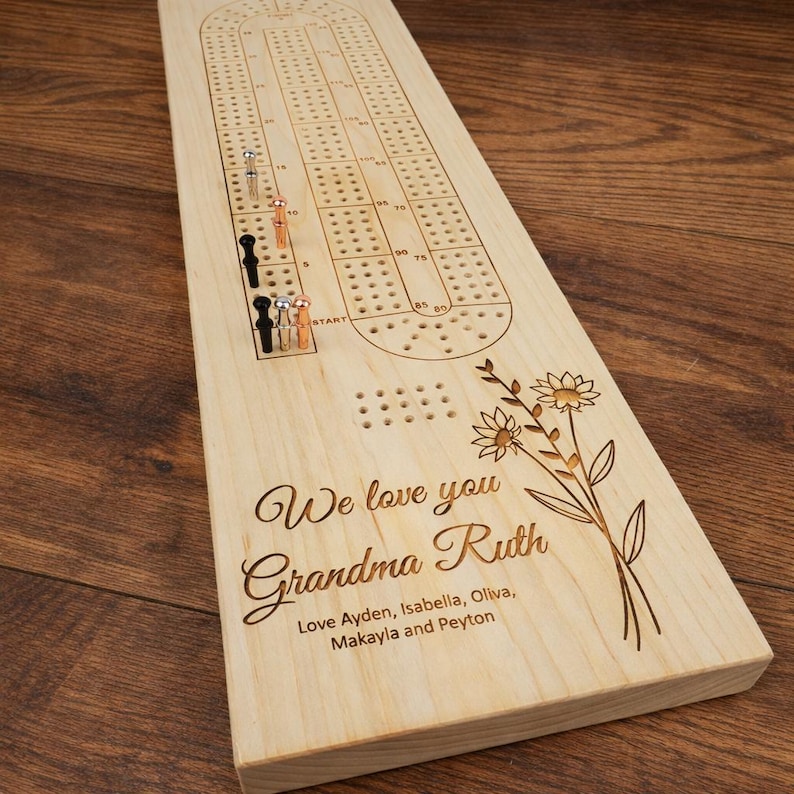 May include: A wooden cribbage board with a floral design and the text "We love you Grandma Ruth". Includes metal pegs in black, silver, and copper colors. The board is made of light-colored wood.
