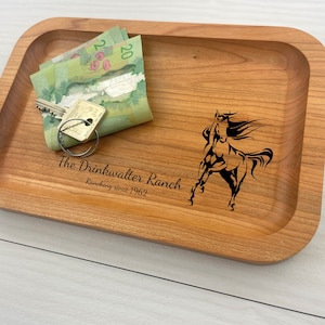 May include: A wooden tray with a horse design and the text "The Drinkwalter Ranch, Ranching since 1962". The tray has a light brown color and is made of wood. There are keys and Canadian money on the tray.